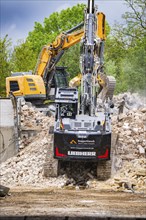 An excavator demolishes a building, surrounded by rubble. Trees and sky in the background,