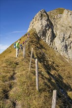 People hiking on a narrow mountain path with a steep ascent and rocky cliffs on a sunny day, saas