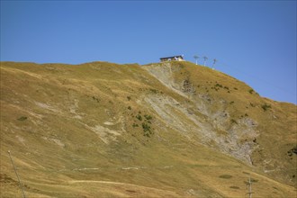 Extensive mountain landscape with a hut on a hill, crossed by a cable car under a blue sky,