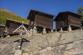 Traditional wooden houses and a cross on a rocky mountainside under a blue sky, Saas Fee,