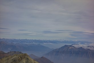 A vast mountain landscape with visible peaks and distant mountain ranges under a cloudy sky, Saas