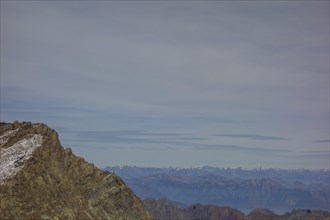 A rocky mountain peak under a cloudy sky with a view of distant mountain ranges, Saas Fee,