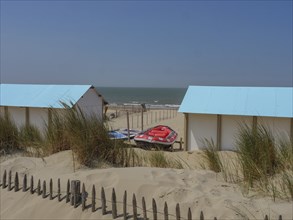 Wooden huts on the beach, surrounded by sand dunes and lifeboats, with the sea in the background,