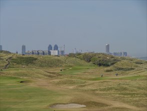 A golf course surrounded by green hills, behind it a city with several tall buildings under a blue