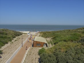 Coastal landscape with wooden house between sand dunes and a path to the beach under a blue sky,