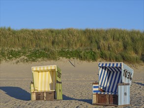 Two coloured beach chairs stand on the sandy beach in front of grassy dunes under a blue sky,