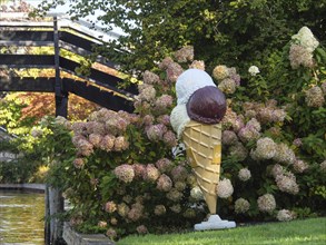 Large ice-cream cone decoration between hydrangea flowers on a canal next to a wooden bridge,