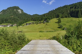 A wooden bridge overlooks green land with several houses, trees and mountains in the background on