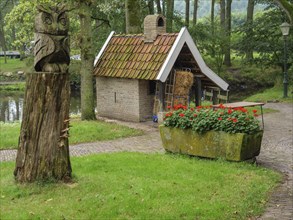 A rural garden with a carved wooden owl, a small brick house, a profusion of flowers and old trees,