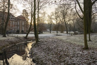 A small stream with a reflection flows through a frosty park, in the background a large building