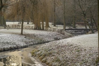 Frost-covered trees and grass surround a small river with a bridge in a wintry park, Velen, North