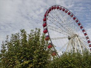 A red and white Ferris wheel towers over green foliage, with a cloudy sky in the background,