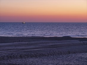 A boat sails on the sea during sunset, the beach shows traces in the sand and the atmosphere is