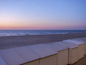 White huts stand on a quiet beach as the sun sets under a purple sky, de haan, Belgium, Europe