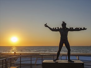 A sculpture on the beach at sunset, the sea in the background. The atmosphere is calm and