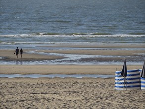 A beach with sand and sea, two people walking along the shore. Blue and white parasols and a quiet