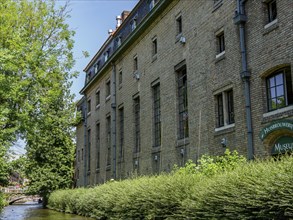 Large historic building along a canal in Bruges, surrounded by trees and green foliage, Bruges,