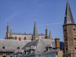 Historic building with many towers and battlements, grey roofs and bricks, under a blue sky,
