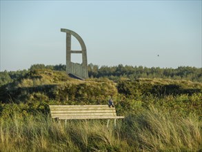 A park bench stands next to a meadow and a sculpture in the background under a clear sky, langeoog,