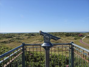 A pair of binoculars on a viewing platform with a view of a vast dune landscape and the sea,