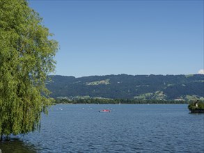 A lake with a red boat and trees in the foreground, mountains in the background under a blue sky,