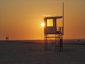 A rescue tower stands on the beach in front of a bright orange sunset, the sea in the background,