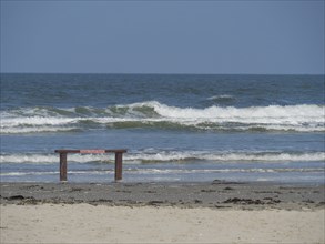 Sea beach with waves and empty bench in the foreground under a bright blue sky, Juist, East Frisia,