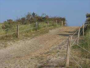 Sandy path leads through the dunes, fenced off by a wooden fence and bright blue sky, juist, east