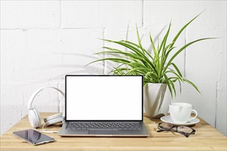 Wooden desk with open laptop mockup, headphones, phone, glasses and plant in front of a white