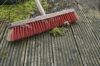 Outdoor broom with red plastic bristles on a weathered wooden deck with slippery algae and moss,