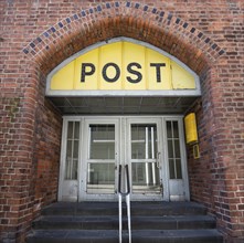 Entrance to the Post office in an historic building with a yellow sign, an old door and an arch of