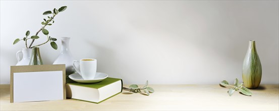 Mock-up of a blank greeting card, vases with sage leaf branch and coffee cup on a book, wooden