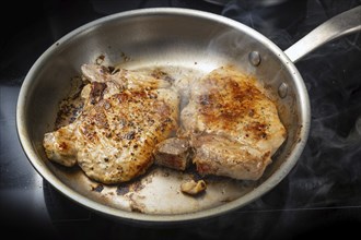 Two pork chops are roasted in a stainless steel frying pan on a black cooktop, food and cooking