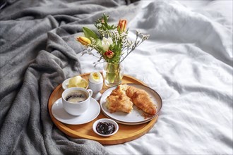 Wooden breakfast tray with croissant, coffee, jam, egg and a fresh flower bouquet served on bed