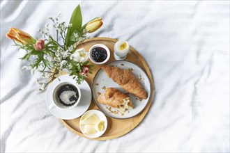 Lazy breakfast in bed, wooden tray with croissant, coffee, jam, egg, butter and flowers on gray