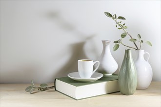 Still live of three vases with a sage leaf branch, gray green book and a coffee cup on a wooden