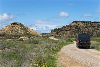 Black van on a gravel road in a hilly, green landscape under a blue sky with clouds, motorhome,