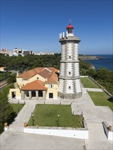 A lighthouse with a red top next to a yellow building, surrounded by a tarmac path and a view of