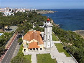 A lighthouse on the coast with a red top and a neighbouring building next to a forest and a paved