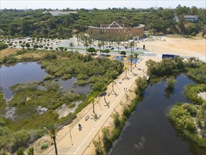 Large park with walking paths, green vegetation, a building in the background and clear sky, aerial