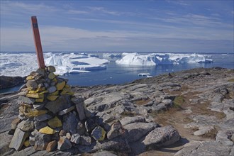 A pile of stones with a flag on rocky terrain in front of an arctic landscape and icebergs in the