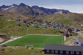 Village with colourful houses and a green football pitch surrounded by mountains and clear skies,