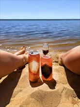 Two people relaxing on a sandy beach with a bottle and can of drink by the water on a sunny day