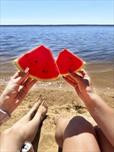 Two hands holding watermelon slices on a sandy beach by a calm lake under a clear sunny sky