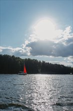 Sailboat with a red sail on a lake under a bright sun and scattered clouds, surrounded by forest