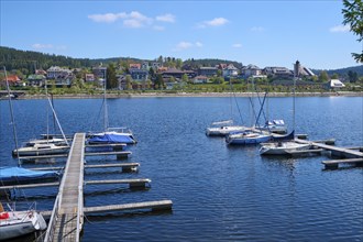 A marina at the Schluchsee with several boats in the water, in the background a town and green