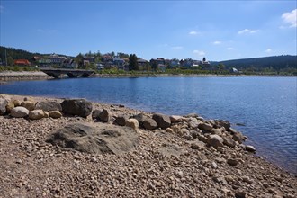 Quiet lake landscape, Schluchsee with pebble beach, bridge and houses on a hill under blue sky,