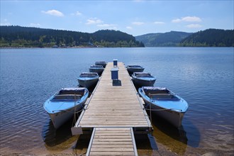 Jetty with moored boats on the Schluchsee, surrounded by mountains and forests, under blue sky,