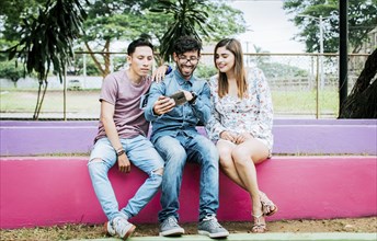 Three happy friends looking at the cell phone sitting in the park. Three happy people looking at