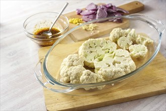 Raw cauliflower slices in a glass casserole, chopped red onion and a spice mix in olive oil,